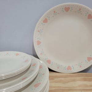 May include: A stack of white ceramic dinner plates featuring a repeating pattern of soft pink hearts and small teal floral accents along the rim. The plates have a smooth, circular design with a consistent decorative border around the edge.