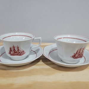 May include: Two white teacups and saucers with a red and white ship design. The cups have a rounded shape and a handle on the right side. The saucers are round with a scalloped edge.