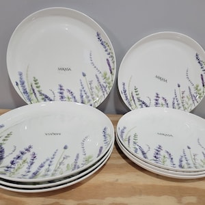 May include: A set of Mikasa white ceramic plates with a lavender floral design. The set includes large round plates and smaller oval plates. The lavender design features purple and green accents. The plates are arranged on a wooden surface.