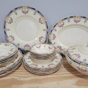 May include: A collection of vintage cream-colored china with floral and blue trim. The set includes plates, bowls, and a serving dish. The plates have a gold rim and the floral design includes pink and yellow flowers.