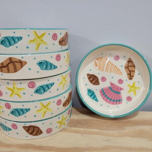 May include: A stack of six ceramic bowls and a single plate, all decorated with a sea life theme. The bowls and plate are white with a teal rim and feature illustrations of seashells, starfish, and dots in various colors.