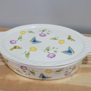 May include: White oval ceramic casserole dish with a lid. The dish and lid are decorated with colorful butterflies, yellow and purple flowers, and green leaves. The dish has two handles and a textured rim.