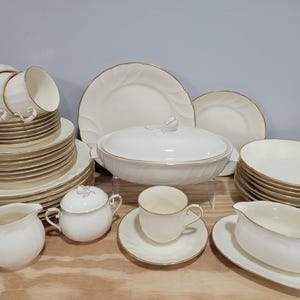 May include: A complete white porcelain dinnerware set with gold trim. The set includes plates, bowls, cups, saucers, a serving dish with a lid, a creamer, a sugar bowl, and a gravy boat. The dishes are stacked and arranged on a wooden surface.