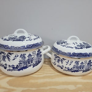 May include: Two white enamel pots with blue and white willow pattern designs. The pots have lids and silver handles.