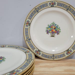 May include: White ceramic plates with a blue and gold decorative border. Each plate features a central image of a basket overflowing with colorful fruits. A stack of plates is visible.
