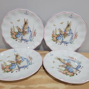 May include: Four white ceramic plates with scalloped edges and pink rims. Each plate features a colorful illustration of two rabbits, flowers, and butterflies. The plates are arranged on a wooden surface.