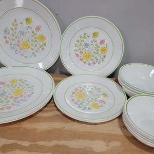 May include: A collection of white ceramic dinnerware with a floral design. The set includes plates of various sizes and bowls. Each piece has a green rim and a central floral pattern with yellow, blue, and pink flowers.