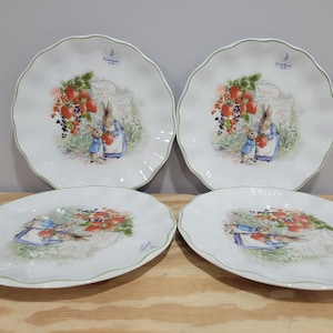 May include: Set of four white ceramic plates with a green scalloped edge. Each plate features a colorful illustration of Peter Rabbit and his friends in a garden setting with strawberries.