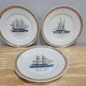 May include: Three white ceramic plates with gold trim and red and blue accents. Each plate features a black and white illustration of a sailing ship with text about the ship.