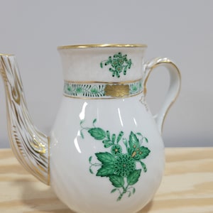 May include: A white porcelain teapot with a green and gold floral design. The teapot has a curved spout and a handle.