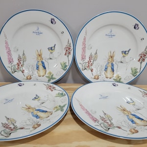 May include: Set of four white ceramic plates with blue trim. Each plate features a Peter Rabbit illustration with a bluebird, flowers, and greenery. The plates are perfect for serving a meal or for decorative purposes.