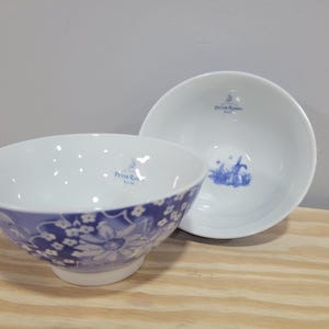 May include: Two white ceramic bowls. One bowl has a blue floral pattern around the rim. The other bowl is plain white with a blue illustration and text. Both bowls have the text "Peter Rabbit" in blue.