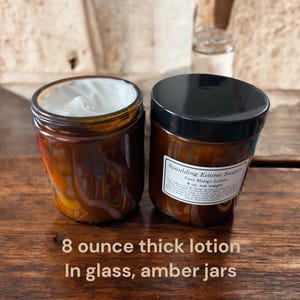 May include: Two amber glass jars filled with thick white lotion. The jars have black lids and are labeled "Spaulding Estates Soapery Coco Mango Lotion 8 oz. net weight".