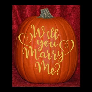 May include: A bright orange pumpkin featuring a green stem and the yellow cursive text Will you Marry Me? with decorative heart-shaped flourishes on the sides. The pumpkin has a smooth, rounded surface with vertical ridges and a natural texture.