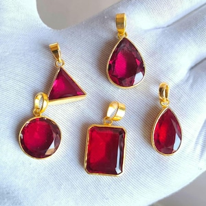 May include: Five gold pendants, each with a different shape: circle, triangle, rectangle, and two teardrops. Each pendant holds a vibrant red gemstone. The pendants are displayed on a white surface.