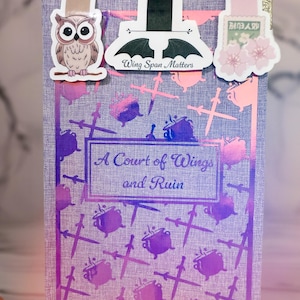 May include: A purple book with a holographic cover featuring a repeating pattern of cauldrons and swords. The book title is "A Court of Wings and Ruin". Three bookmarks are attached to the book: an owl, a bat with the text "Wing Span Matters", and a pink flower with Japanese text.