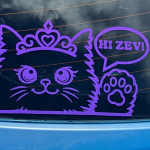 Könnte beinhalten: Ein violetter Glitzer-Vinylaufkleber, der eine flauschige Katze mit Tiara zeigt, die eine Pfote hebt, neben einer Sprechblase mit dem Text HI ZEV!. Das Design ist für die Anbringung auf glatten Oberflächen wie Autoscheiben vorgesehen.