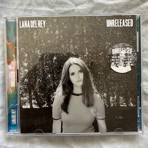 Peut inclure: Une photo en noir et blanc d'une femme aux cheveux longs, portant un chemisier rayé rouge et blanc. La photo est sur un boîtier de CD avec le texte "LANA DEL REY" et "UNRELEASED" dessus.