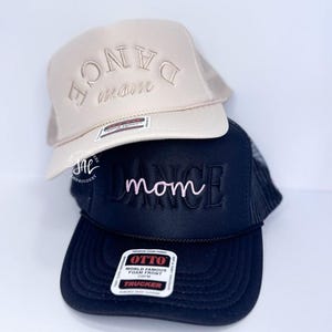May include: Two trucker hats stacked on top of each other. The top hat is tan with "DANCE mom" embroidered in a matching color. The bottom hat is black with "DANCE mom" embroidered in pink.