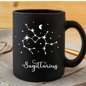 Puede incluir: Taza de caf&eacute; de cer&aacute;mica negra con un dise&ntilde;o blanco de la constelaci&oacute;n de Sagitario y la palabra "Sagitario" en una fuente cursiva.