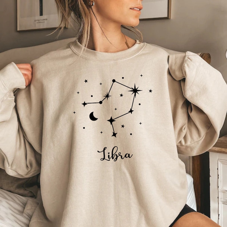 Puede incluir: Una sudadera beige con un gr&aacute;fico negro de la constelaci&oacute;n zodiacal de Libra y la palabra "Libra" impresa debajo.