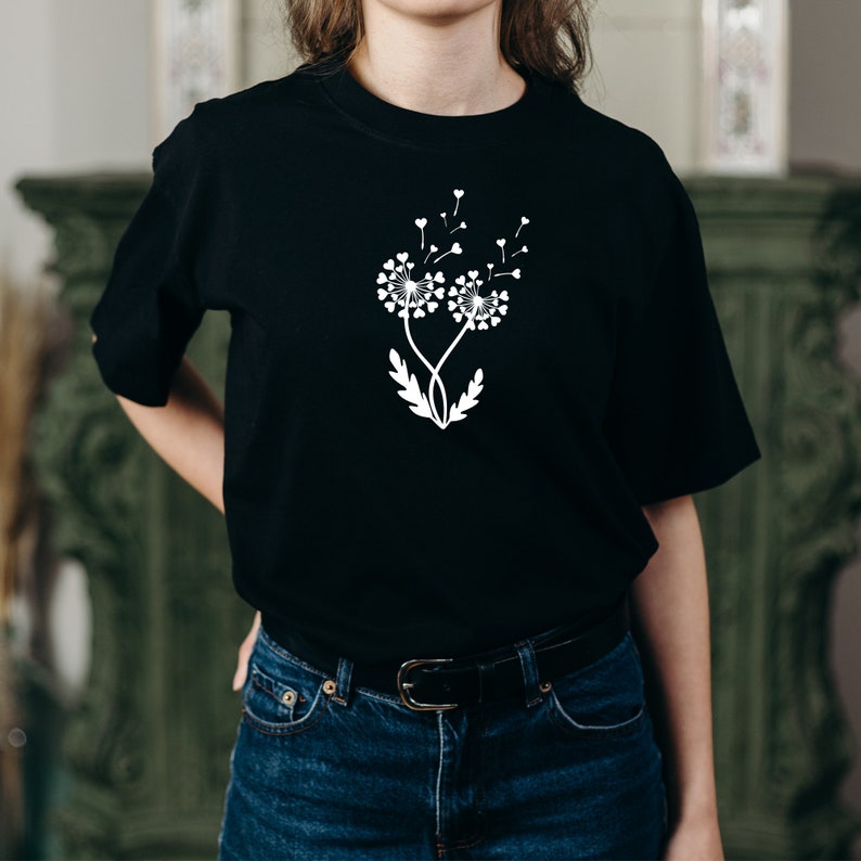 K&ouml;nnte beinhalten: Ein schwarzes T-Shirt mit einem wei&szlig;en Design aus zwei L&ouml;wenzahnpflanzen mit Herzen als Samen. Die L&ouml;wenz&auml;hne werden vom Wind verweht.