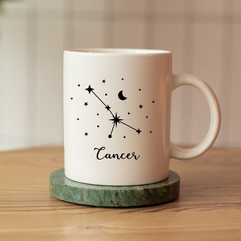 Puede incluir: Taza de cer&aacute;mica blanca con un dise&ntilde;o de constelaci&oacute;n del zod&iacute;aco C&aacute;ncer negro y la palabra "C&aacute;ncer" impresa debajo. La taza est&aacute; sobre un posavasos de m&aacute;rmol verde.
