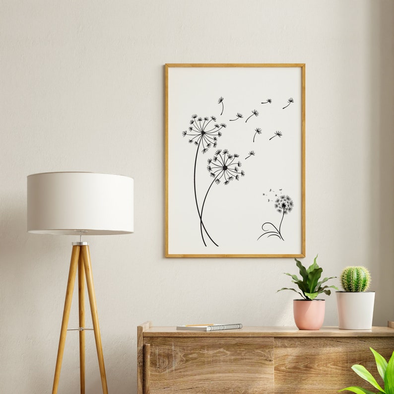 K&ouml;nnte beinhalten: Eine Schwarzwei&szlig;illustration von L&ouml;wenzahnpflanzen mit Samen, die im Wind fliegen, gerahmt in einem hellen Holzrahmen und an einer wei&szlig;en Wand &uuml;ber einer Kommode aus Holz aufgeh&auml;ngt.