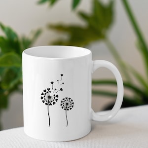 K&ouml;nnte beinhalten: Wei&szlig;e Keramiktasse mit einer schwarzen Silhouette von zwei L&ouml;wenzahnpflanzen mit Herzen als Samen. Der gr&ouml;&szlig;ere L&ouml;wenzahn hat einige Samen, die wegfliegen.