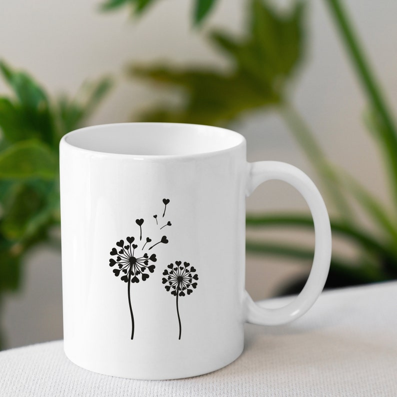 K&ouml;nnte beinhalten: Wei&szlig;e Keramiktasse mit einer schwarzen Silhouette von zwei L&ouml;wenzahnpflanzen mit Herzen als Samen. Der gr&ouml;&szlig;ere L&ouml;wenzahn hat einige Samen, die wegfliegen.