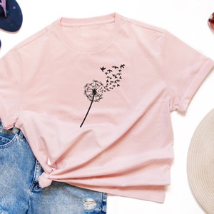 K&ouml;nnte beinhalten: Ein rosa T-Shirt mit einem schwarzen L&ouml;wenzahnmuster und einer Vogelschar, die vom L&ouml;wenzahn wegfliegt.