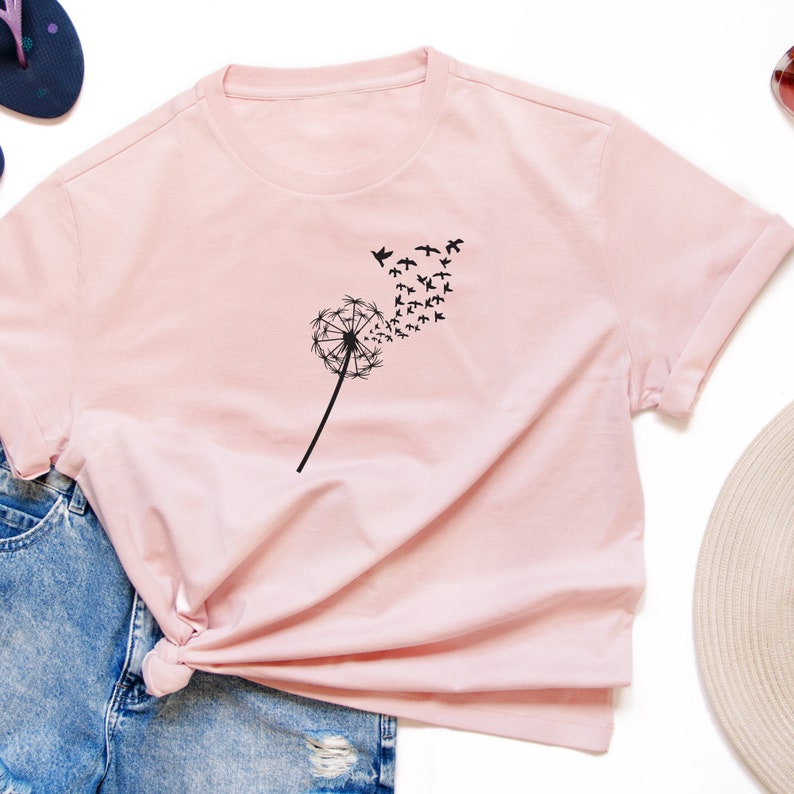 K&ouml;nnte beinhalten: Ein rosa T-Shirt mit einem schwarzen L&ouml;wenzahnmuster und einer Vogelschar, die vom L&ouml;wenzahn wegfliegt.