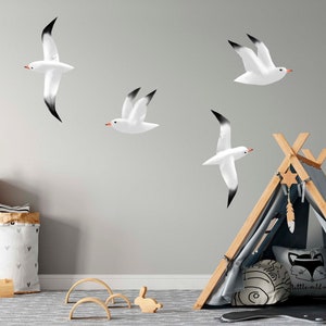 Peut inclure: Quatre autocollants muraux en forme de mouette blanche avec des extrémités d'ailes noires, sur un mur gris clair. Les autocollants sont disposés en quinconce. Une tente tipi grise et blanche avec une armature en bois se trouve dans le coin inférieur droit de l'image.
