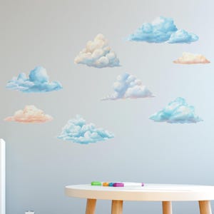 May include: A collection of cloud wall decals in various sizes and colors, including light blue, peach, and white. The decals are arranged on a light gray wall, creating a whimsical and airy atmosphere. A small, round table with colorful markers is in the foreground.