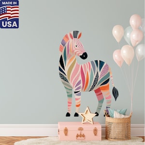 Puede incluir: Un vinilo decorativo de pared con un cebra colorida con melena y rayas corporales arcoíris. La cebra está junto a un baúl rosa, una decoración en forma de estrella y una cesta de almohadas. Globos atados. Hecho en los EE. UU.