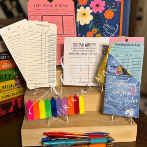 May include: A tiered wooden stand with a variety of colorful items, including a reading log, a "You Said What?!" game card, a travel book log, a denim pouch with the text "Sisterhood of the Traveling Books", and a stack of colorful pens. The stand is decorated with rainbow-colored sticky notes.