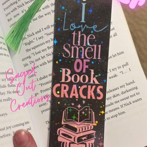 May include: Black bookmark with the text "I Love the Smell of Book Cracks" in pink and blue. The bookmark has a stack of books graphic and a green tassel. The word "BEST SELLER" is in the top right corner.