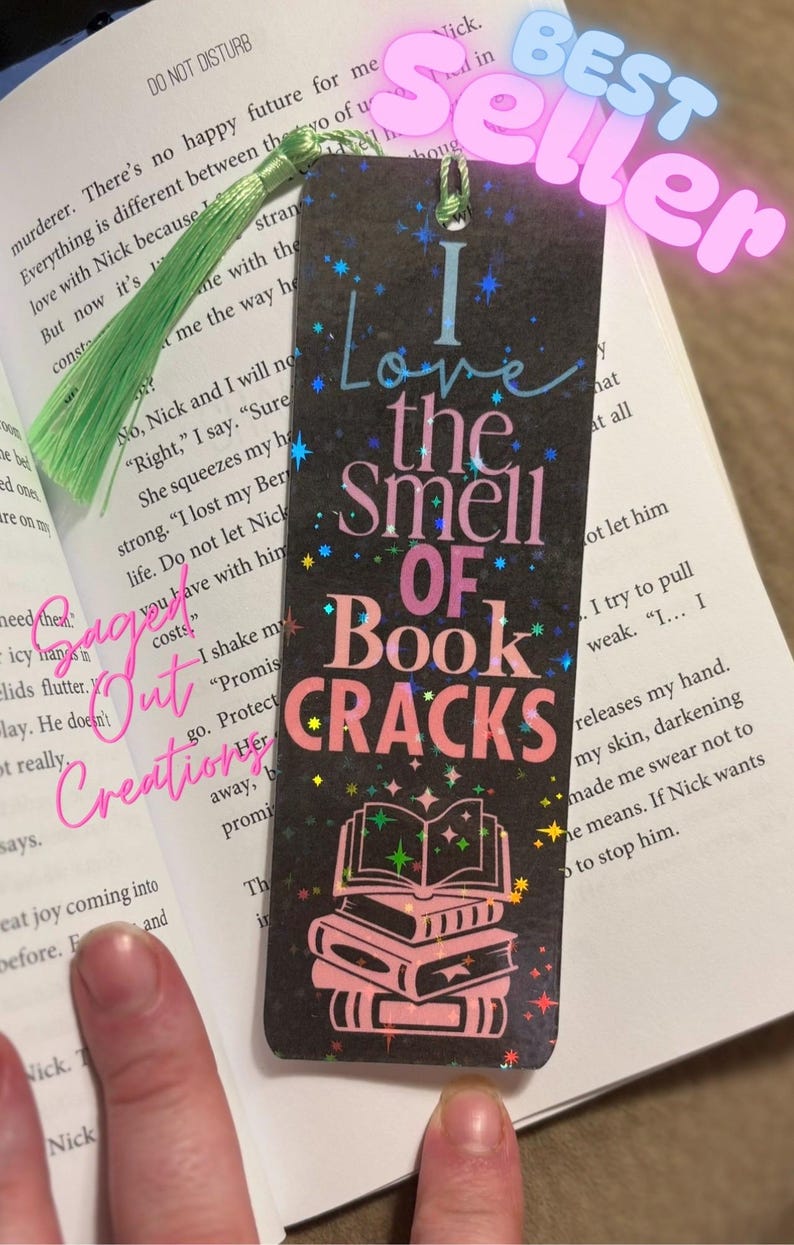 May include: Black bookmark with the text "I Love the Smell of Book Cracks" in pink and blue. The bookmark has a stack of books graphic and a green tassel. The word "BEST SELLER" is in the top right corner.