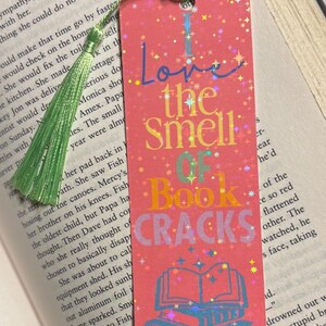 May include: Pink bookmark with the text "Love the Smell of Book Cracks" in colorful letters. The bookmark has a stack of books graphic and a green tassel.