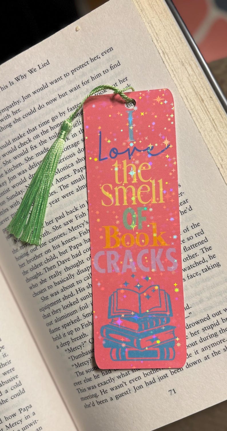 May include: Pink bookmark with the text "Love the Smell of Book Cracks" in colorful letters. The bookmark has a stack of books graphic and a green tassel.