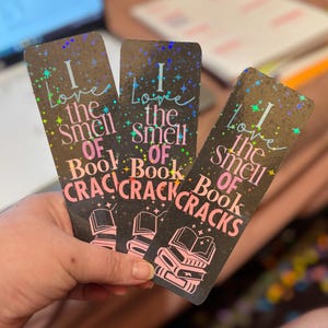 May include: Three black bookmarks with holographic accents are held in a hand. Each bookmark reads "I love the smell of book cracks" in pink and white text, with a stack of books graphic.