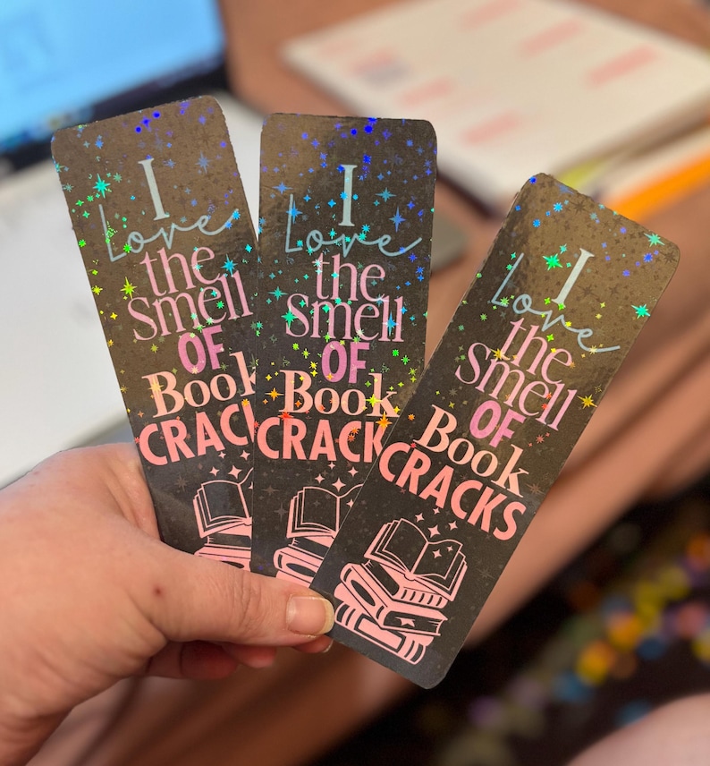 May include: Three black bookmarks with holographic accents are held in a hand. Each bookmark reads "I love the smell of book cracks" in pink and white text, with a stack of books graphic.