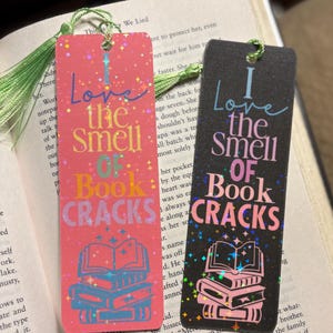 May include: Two rectangular bookmarks with the text "I Love the Smell of Book Cracks." One is pink with blue and orange accents, the other is black with pink accents. Both have a tassel and a stack of books graphic.