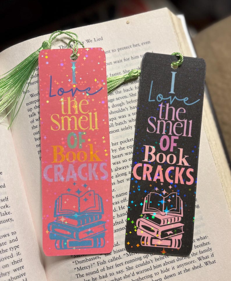 May include: Two rectangular bookmarks with the text "I Love the Smell of Book Cracks." One is pink with blue and orange accents, the other is black with pink accents. Both have a tassel and a stack of books graphic.