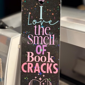 May include: A rectangular bookmark with the text "I love the smell of book cracks" in various colors. The bookmark has a black background with colorful star accents and a pink ribbon attached at the top. A stack of books is illustrated at the bottom.
