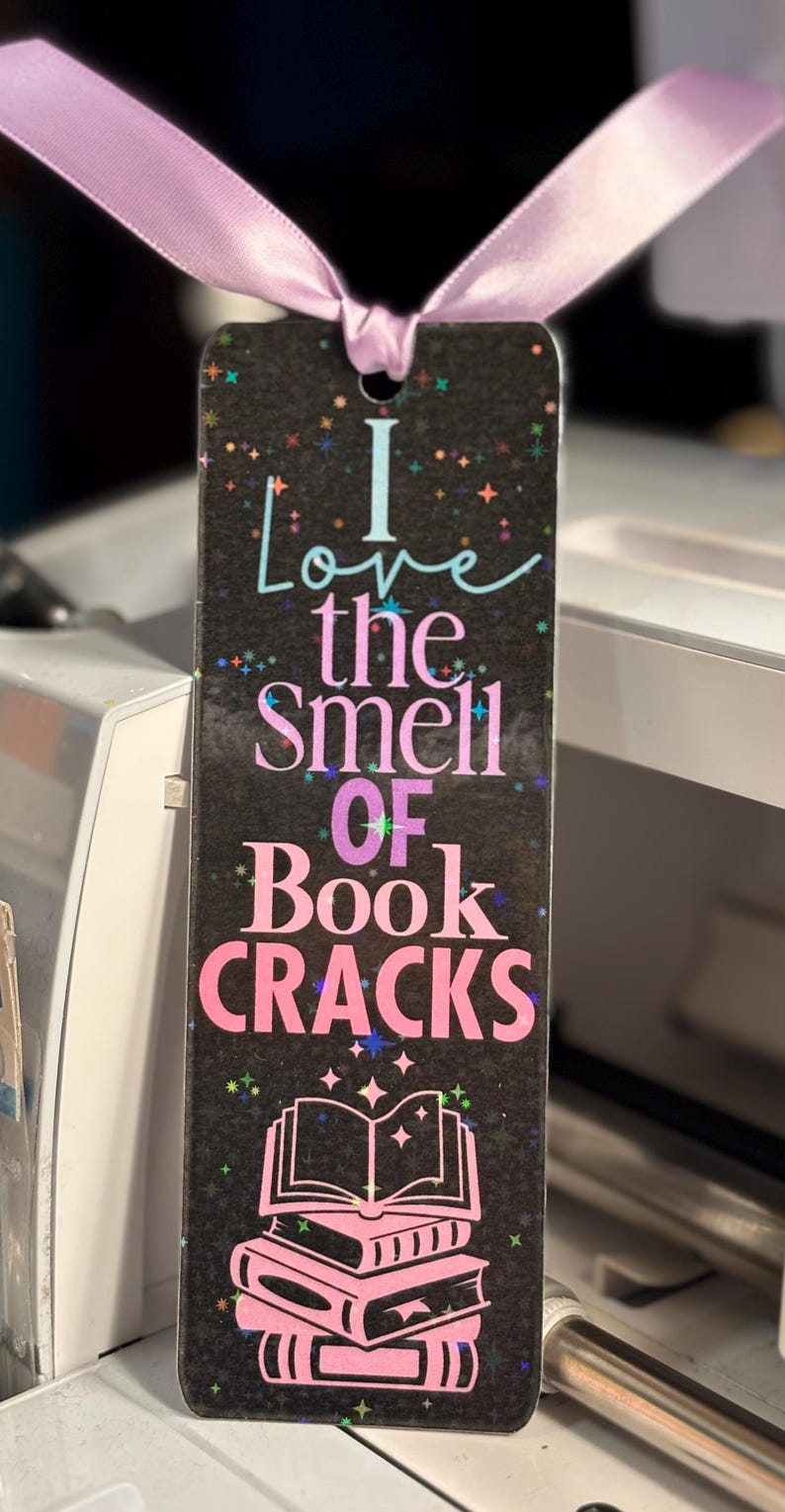 May include: A rectangular bookmark with the text "I love the smell of book cracks" in various colors. The bookmark has a black background with colorful star accents and a pink ribbon attached at the top. A stack of books is illustrated at the bottom.