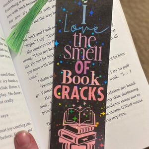 May include: A black bookmark with the text "I Love the Smell of Book Cracks" in colorful lettering. The bookmark has a green tassel and a stack of pink books illustration. It is placed in an open book.