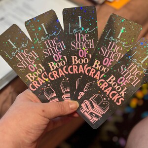 May include: A hand holds five rectangular bookmarks. Each bookmark has a dark background with the text "I love the smell of book cracks" in white and pink. The bookmarks have a holographic, glittery effect.