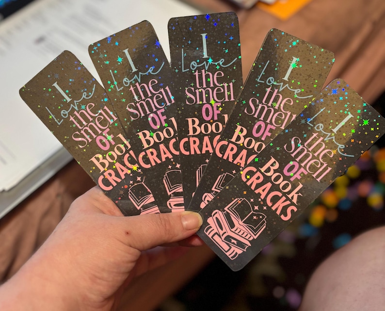 May include: A hand holds five rectangular bookmarks. Each bookmark has a dark background with the text "I love the smell of book cracks" in white and pink. The bookmarks have a holographic, glittery effect.