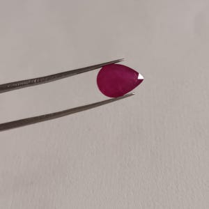 May include: A pear-shaped, deep red gemstone held by tweezers against a white background.
